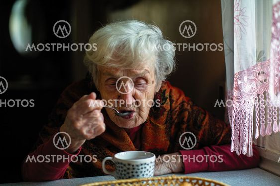 An elderly retired woman drinking tea from a spoon,...
