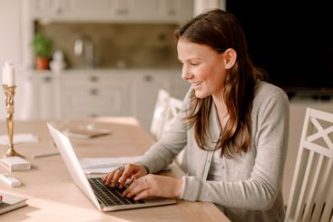 Smiling teenage girl doing homework while using laptop at...