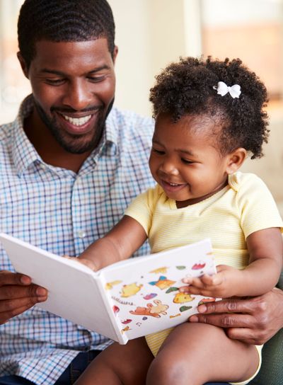 Baby Daughter And Father Sitting On Sofa Reading Book...