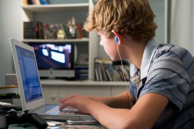 Young boy in bedroom using laptop and listening to MP3...