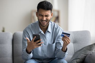 Excited millennial male sit on sofa hold phone credit card