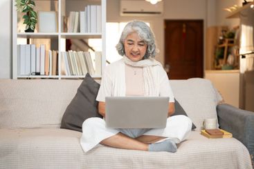 A happy Asian senior woman uses a laptop while sitting...