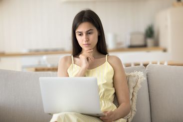 Serious young woman sitting on couch reading email on...