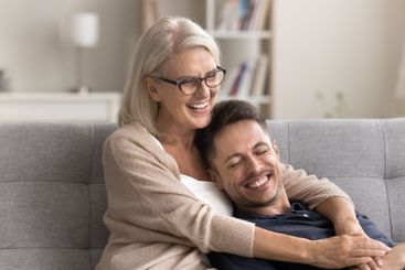 Grownup son relax on couch in loving senior mommy embraces