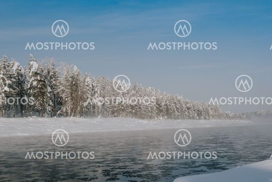Frosty fog over winter river with snow and forest on...